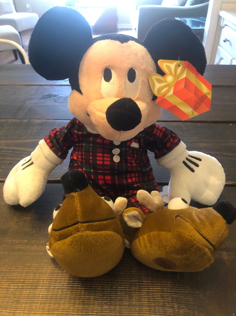 May include: A plush Mickey Mouse wearing red and black plaid pajamas and reindeer slippers. The plush toy is holding a gift box with a yellow bow.