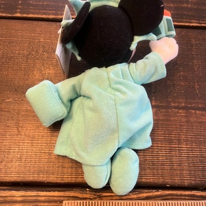 May include: A plush toy of Mickey Mouse dressed as the Statue of Liberty. He is wearing a green robe with a crown on his head. He is holding a torch in his right hand.
