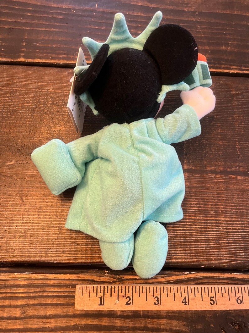 May include: A plush toy of Mickey Mouse dressed as the Statue of Liberty. He is wearing a green robe with a crown on his head. He is holding a torch in his right hand.
