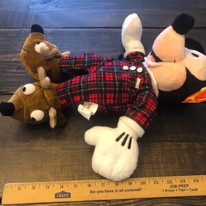 May include: A red and black plaid plush Mickey Mouse wearing a pair of brown reindeer slippers. The Mickey Mouse plush is lying on a wooden surface.