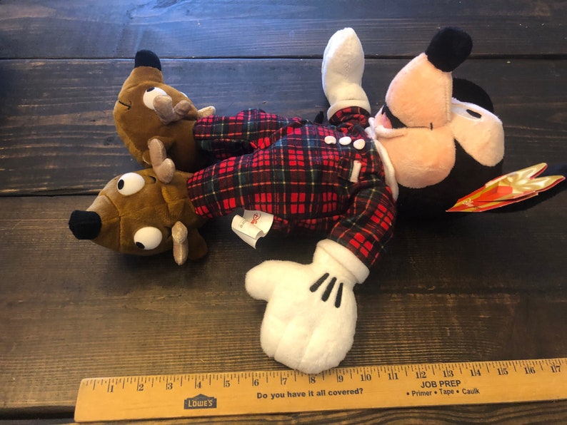 May include: A red and black plaid plush Mickey Mouse wearing a pair of brown reindeer slippers. The Mickey Mouse plush is lying on a wooden surface.