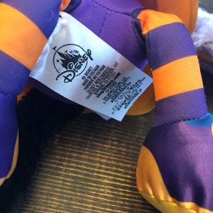 May include: A close-up of a Disney plush toy. The toy has purple and orange striped legs and a yellow foot. A white tag with the Disney logo and text is visible. The plush toy is on a wooden surface.