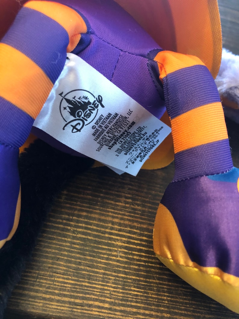 May include: A close-up of a Disney plush toy. The toy has purple and orange striped legs and a yellow foot. A white tag with the Disney logo and text is visible. The plush toy is on a wooden surface.