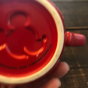 May include: Red ceramic mug with a white interior and a small, raised, circular design on the bottom. The mug has a handle and the text "Dishwasher Safe Microwave Safe" is visible on the bottom.