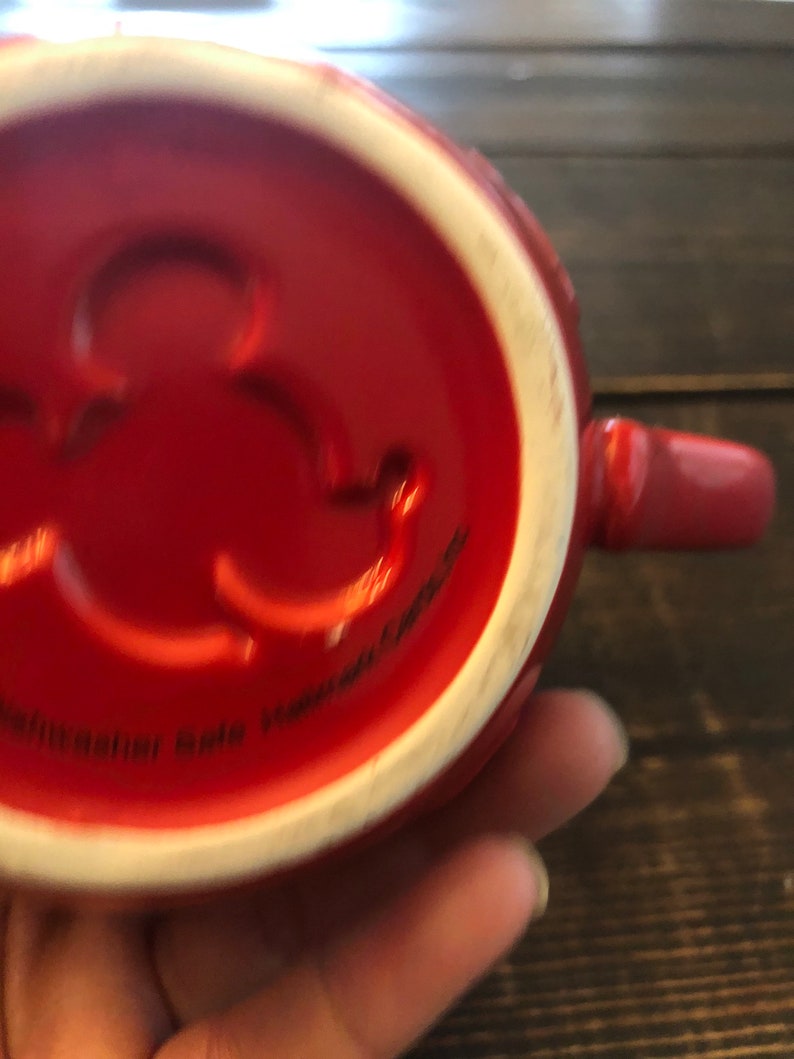 May include: Red ceramic mug with a white interior and a small, raised, circular design on the bottom. The mug has a handle and the text "Dishwasher Safe Microwave Safe" is visible on the bottom.