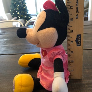 May include: A pink and black plush Minnie Mouse toy wearing a pink dress with a red heart on the front. The toy is sitting on a wooden surface and is about 18 inches tall.