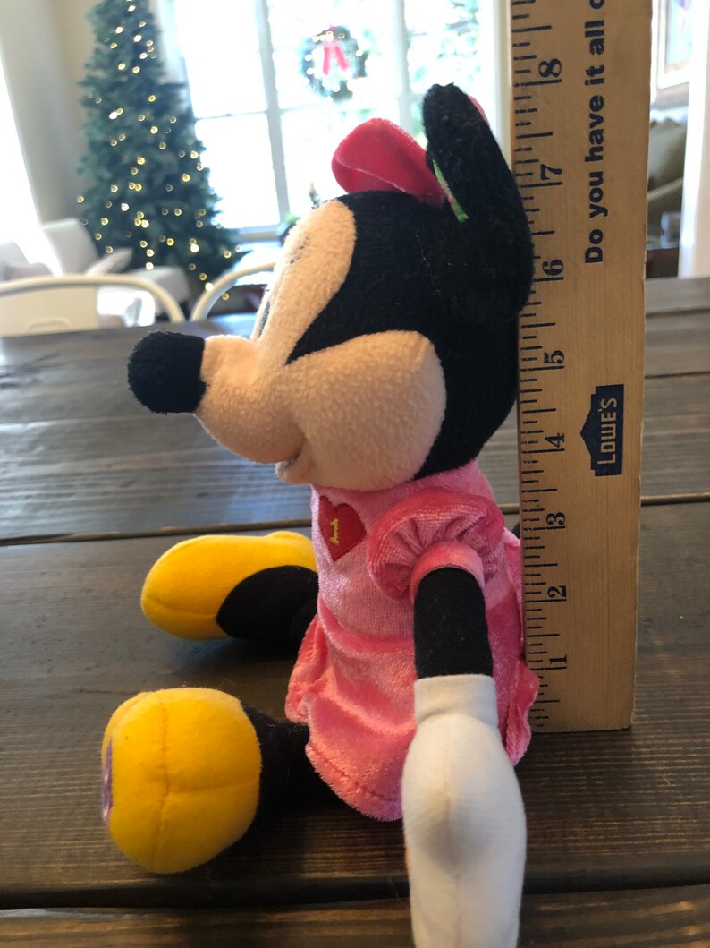 May include: A pink and black plush Minnie Mouse toy wearing a pink dress with a red heart on the front. The toy is sitting on a wooden surface and is about 18 inches tall.