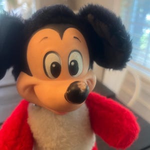 May include: A vintage Mickey Mouse plush toy. The toy features a black head with large ears, a tan face, and a black nose. The body is red with a white chest. The plush toy is a classic Disney character.