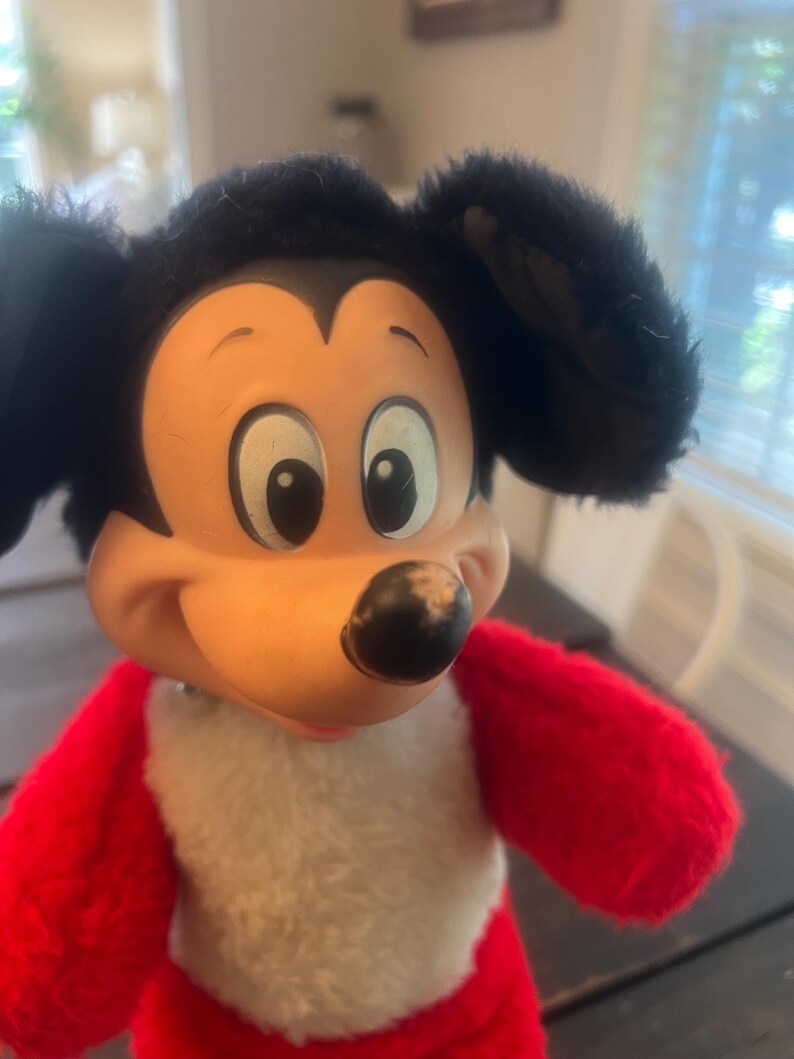 May include: A vintage Mickey Mouse plush toy. The toy features a black head with large ears, a tan face, and a black nose. The body is red with a white chest. The plush toy is a classic Disney character.