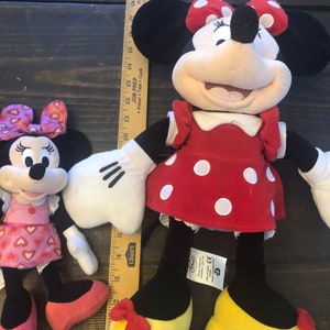 May include: Two plush Mickey Mouse toys. The larger toy is red with white polka dots and a red bow. The smaller toy is pink with white hearts and a pink bow. Both toys have black shoes and white gloves.