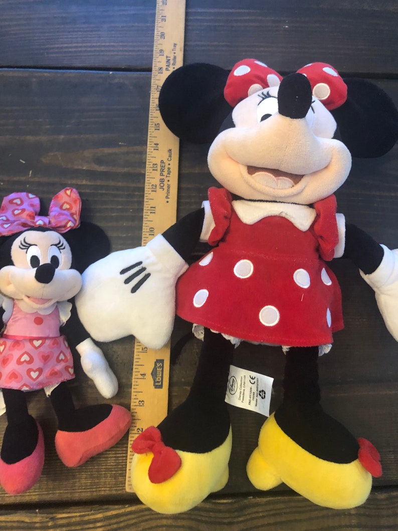 May include: Two plush Mickey Mouse toys. The larger toy is red with white polka dots and a red bow. The smaller toy is pink with white hearts and a pink bow. Both toys have black shoes and white gloves.