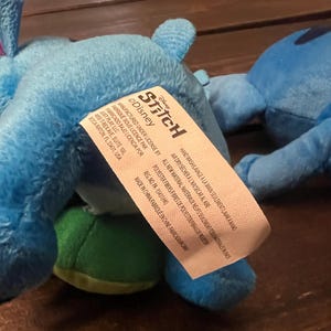 Op de afbeelding: Twee blauwe pluche Stitch-speelgoed, een met een groene buik, worden getoond. Het speelgoed heeft het Disney Stitch-label. Het label bevat tekst in het Engels en Frans.