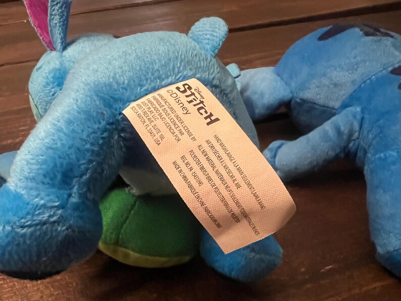 Op de afbeelding: Twee blauwe pluche Stitch-speelgoed, een met een groene buik, worden getoond. Het speelgoed heeft het Disney Stitch-label. Het label bevat tekst in het Engels en Frans.
