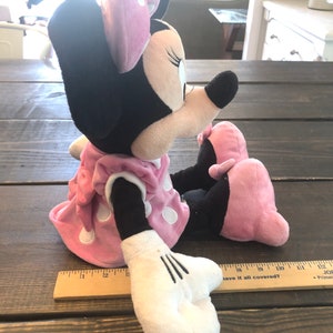 May include: A plush Minnie Mouse toy wearing a pink dress with white polka dots and a pink bow. The toy is sitting on a wooden surface.