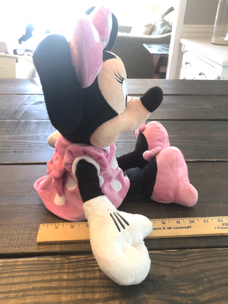 May include: A plush Minnie Mouse toy wearing a pink dress with white polka dots and a pink bow. The toy is sitting on a wooden surface.