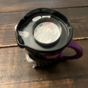 May include: A black and white ceramic mug with a purple handle. The mug features a character design with a black top and a white face. The interior of the mug is black, and the handle is purple.
