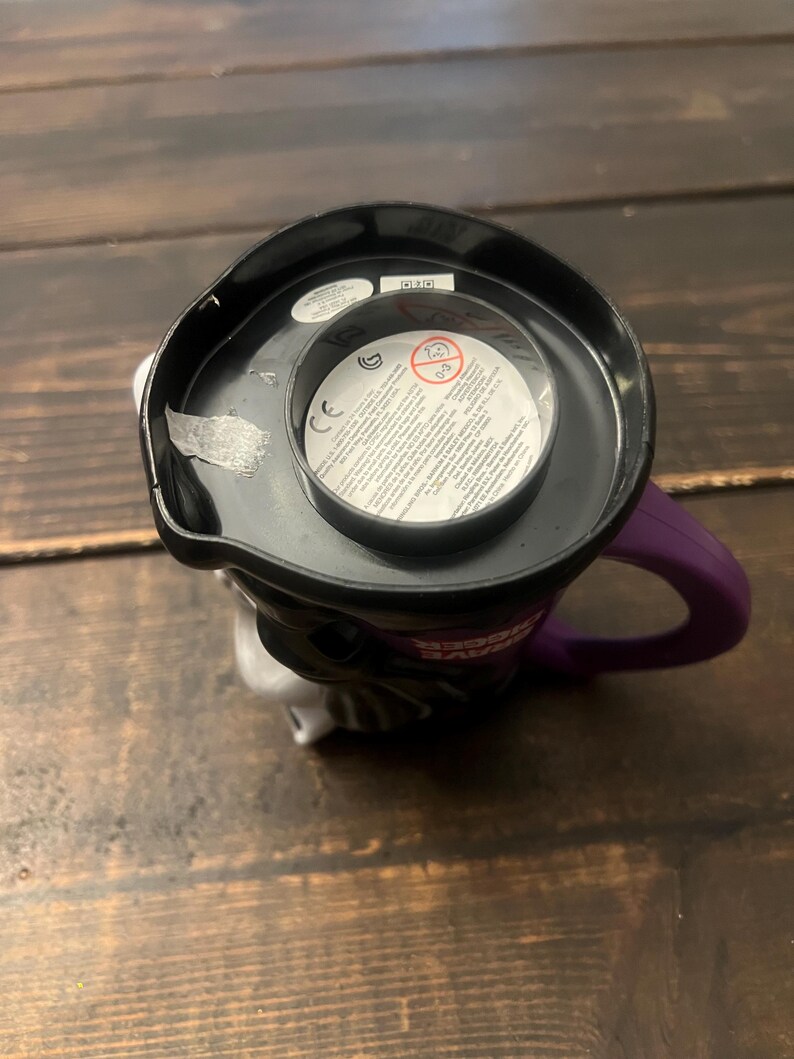 May include: A black and white ceramic mug with a purple handle. The mug features a character design with a black top and a white face. The interior of the mug is black, and the handle is purple.