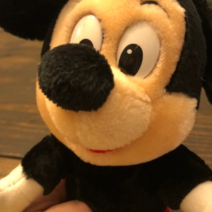 Puede incluir: Un juguete de peluche Mickey Mouse negro y blanco con una pajarita roja y blanca. El juguete tiene una gran sonrisa y ojos grandes.