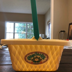 May include: A yellow plastic picnic basket with a green handle and a Cabbage Patch Kids logo. The basket has a diamond pattern.