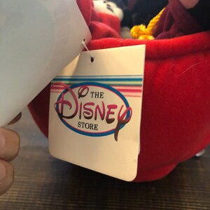 Peut inclure: Une tasse &agrave; th&eacute; en peluche rouge avec une poign&eacute;e en corde jaune. La tasse est remplie de personnages en peluche, dont Mickey Mouse, Minnie Mouse et Dingo. La tasse a une &eacute;tiquette blanche attach&eacute;e &agrave; elle qui indique "The Disney Store".