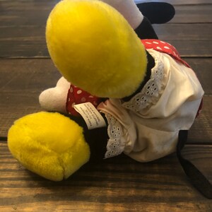 May include: A plush toy of a cartoon character with yellow and black fur, wearing a red and white polka dot dress with a white lace trim. The toy has a black tail and is lying on a wooden surface. The tag on the toy reads "All New Material, Polyester Fiber Filling, Product of Korea."