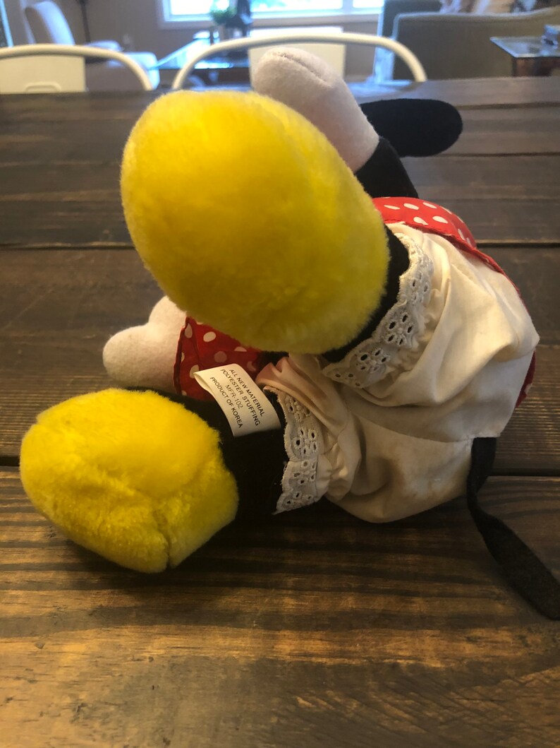 May include: A plush toy of a cartoon character with yellow and black fur, wearing a red and white polka dot dress with a white lace trim. The toy has a black tail and is lying on a wooden surface. The tag on the toy reads "All New Material, Polyester Fiber Filling, Product of Korea."