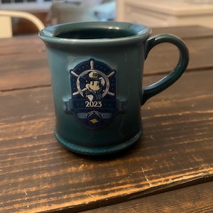 May include: Teal ceramic mug with a handle. The mug features a navy blue emblem with Mickey Mouse in a captain's hat, a ship's wheel, and the year "2023". The mug has a rounded base and a slightly flared rim.