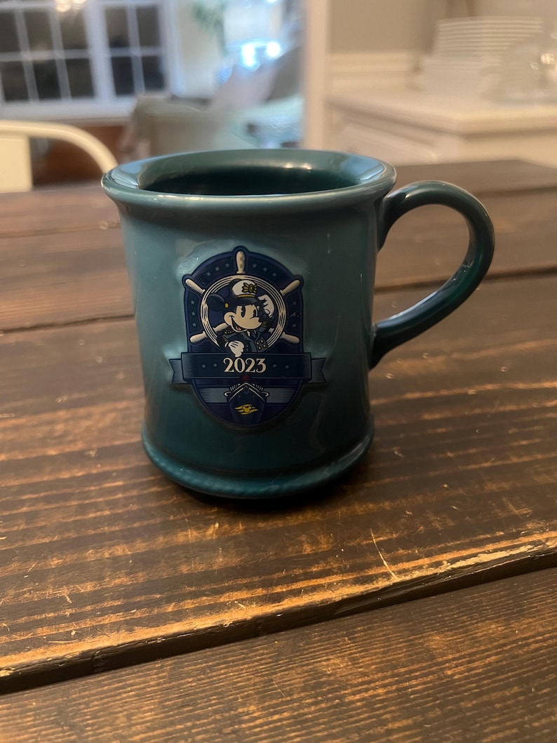 May include: Teal ceramic mug with a handle. The mug features a navy blue emblem with Mickey Mouse in a captain's hat, a ship's wheel, and the year "2023". The mug has a rounded base and a slightly flared rim.