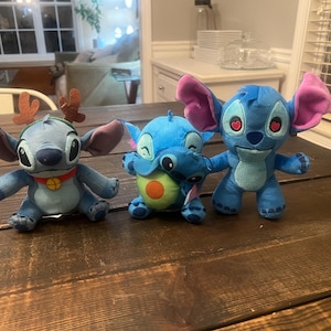 Op de afbeelding: Drie blauwe Stitch-knuffels met grote oren. E&eacute;n draagt een rendiergewei en een rode halsband. Een ander houdt een groene en oranje avocado vast. De derde heeft hartvormige ogen. Ze zitten op een donkere houten ondergrond.