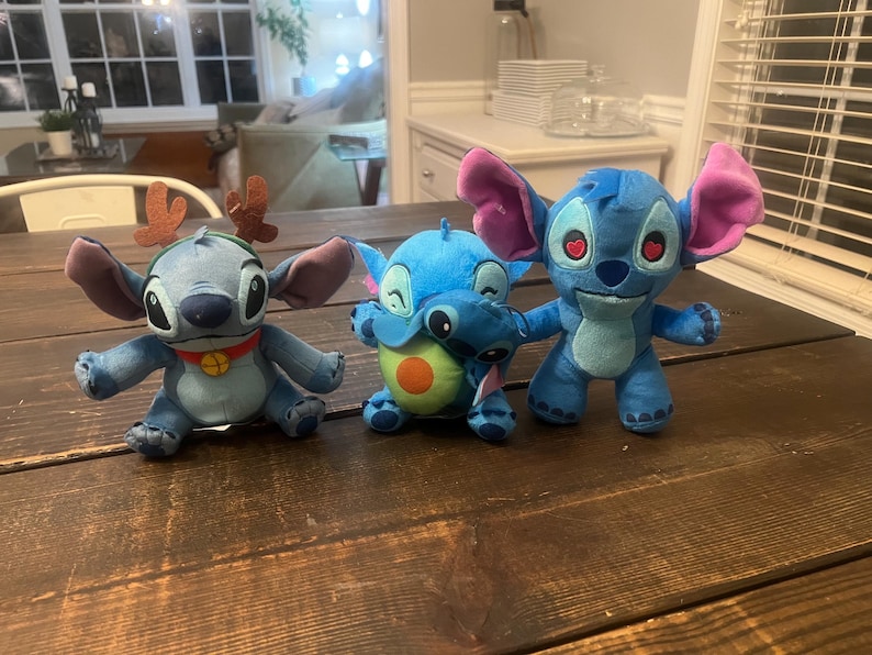Op de afbeelding: Drie blauwe Stitch-knuffels met grote oren. E&eacute;n draagt een rendiergewei en een rode halsband. Een ander houdt een groene en oranje avocado vast. De derde heeft hartvormige ogen. Ze zitten op een donkere houten ondergrond.