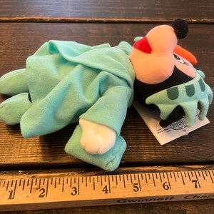 May include: A plush toy of Mickey Mouse dressed as the Statue of Liberty. The toy is wearing a green robe with a crown and is lying on a wooden surface. The toy is approximately 6 inches long.