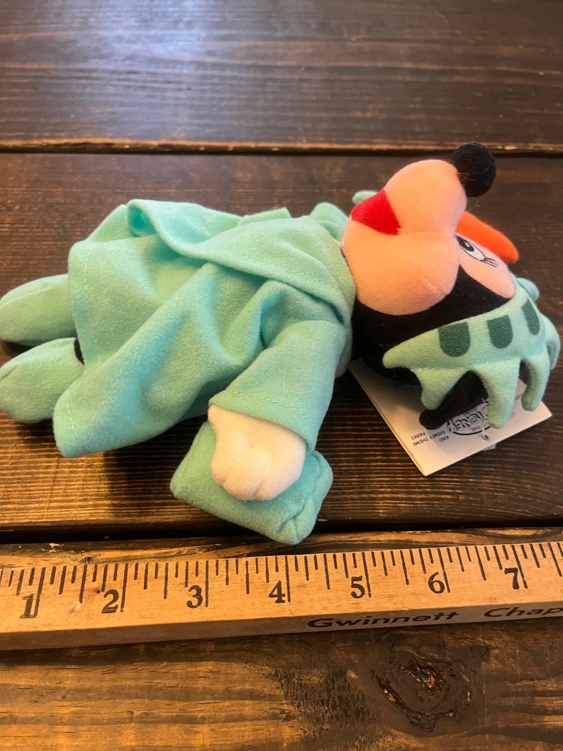 May include: A plush toy of Mickey Mouse dressed as the Statue of Liberty. The toy is wearing a green robe with a crown and is lying on a wooden surface. The toy is approximately 6 inches long.