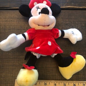 Op de afbeelding: Een pluche rood en zwart Minnie Mouse speelgoed met gele schoenen en een rode strik. Het speelgoed is ongeveer 30 cm hoog en staat op een houten oppervlak.