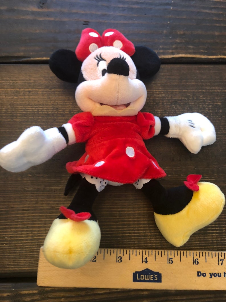 Op de afbeelding: Een pluche rood en zwart Minnie Mouse speelgoed met gele schoenen en een rode strik. Het speelgoed is ongeveer 30 cm hoog en staat op een houten oppervlak.