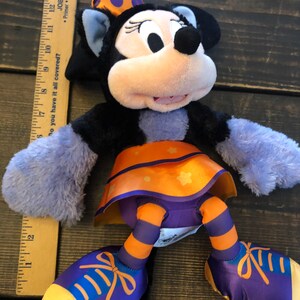 May include: A Minnie Mouse plush toy in a Halloween costume. The plush has a black body, lavender arms, and orange and purple accents. It wears a polka-dot bow and a skirt with star details. A wooden ruler is next to the toy.