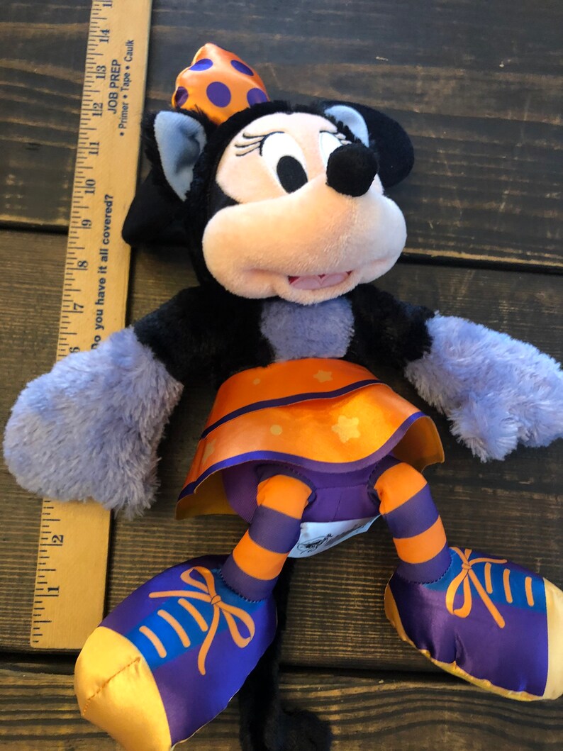 May include: A Minnie Mouse plush toy in a Halloween costume. The plush has a black body, lavender arms, and orange and purple accents. It wears a polka-dot bow and a skirt with star details. A wooden ruler is next to the toy.