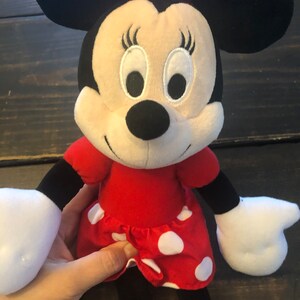 May include: A plush toy of Minnie Mouse wearing a red dress with white polka dots and a red bow. She has black ears, a black nose, and white eyes.