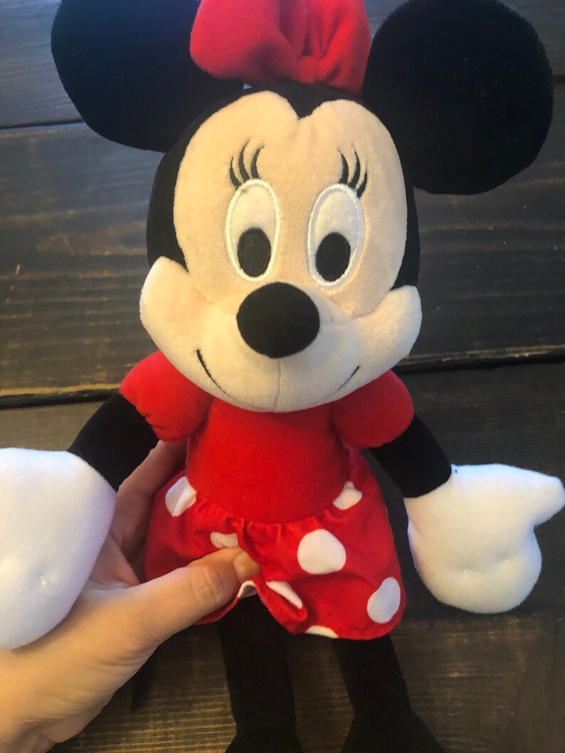 May include: A plush toy of Minnie Mouse wearing a red dress with white polka dots and a red bow. She has black ears, a black nose, and white eyes.