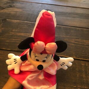 May include: A pink and black Minnie Mouse hand puppet wearing a pink dress with white lace trim and a pink bow.