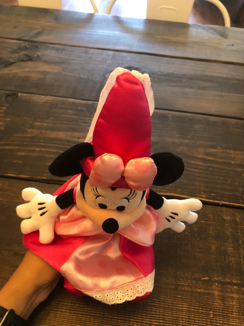 May include: A pink and black Minnie Mouse hand puppet wearing a pink dress with white lace trim and a pink bow.