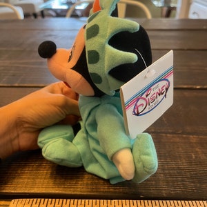 May include: A green plush Mickey Mouse wearing a Statue of Liberty hat. The plush is sitting on a wooden surface with a ruler in the foreground. The tag attached to the plush reads "The Disney Store".