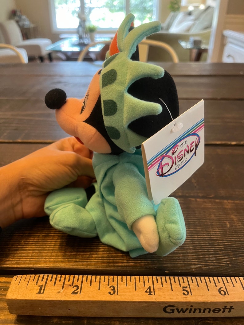 May include: A green plush Mickey Mouse wearing a Statue of Liberty hat. The plush is sitting on a wooden surface with a ruler in the foreground. The tag attached to the plush reads "The Disney Store".