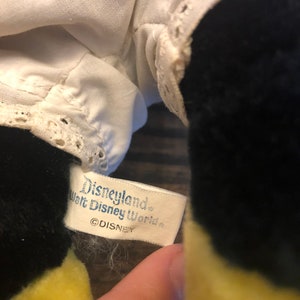 K&ouml;nnte beinhalten: Ein wei&szlig;es Etikett mit schwarzem Text, auf dem "Disneyland Walt Disney World &copy;DISNEY" steht, das an einem schwarzen und gelben Pl&uuml;schtier befestigt ist.