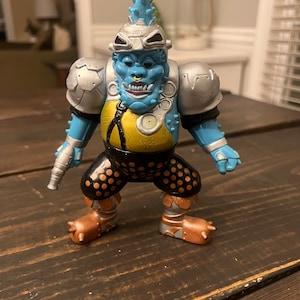 May include: A blue and silver action figure with a spiked mohawk, goggles, and a yellow and black patterned outfit. The figure is holding a silver gun in its right hand.