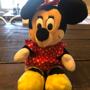 K&ouml;nnte beinhalten: Ein Pl&uuml;schtier von Minnie Mouse, das ein rotes Kleid mit wei&szlig;en Punkten und eine rote Schleife tr&auml;gt. Sie hat schwarze Ohren und eine schwarze Nase, und ihre Augen sind gro&szlig; und wei&szlig;.