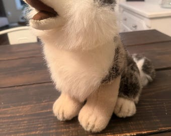 May include: A plush wolf toy with soft, multi-toned fur in shades of grey, white, and tan. The stuffed animal is depicted in a seated position with its head tilted upward, featuring a dark nose, dark eyes, and an open mouth showing a brown interior.