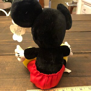 May include: A black and red plush Mickey Mouse toy sitting on a wooden surface. The toy is approximately 13 inches tall. The toy has a red and white tag attached to its ear that reads "Celebrate the magic of Disney and the joy of giving.  This plush toy is perfect for cuddling and sharing the love of Mickey Mouse.  Made with high-quality materials and designed for lasting fun.  Mickey Mouse is a registered trademark of The Walt Disney Company."