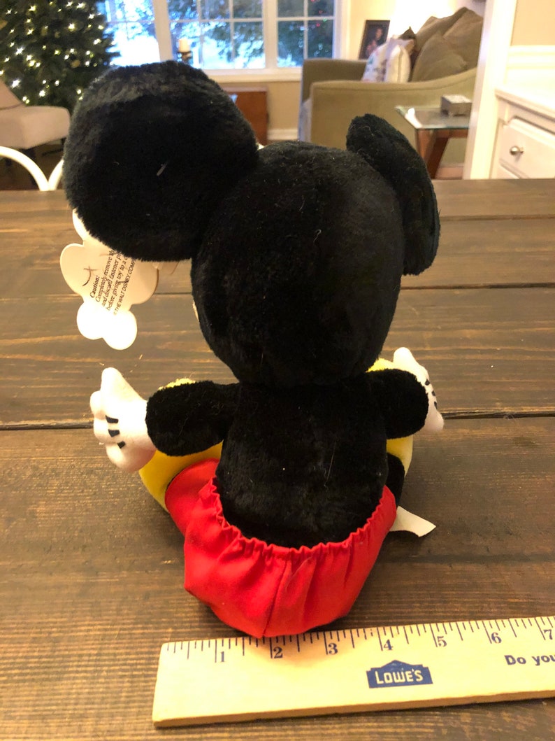 May include: A black and red plush Mickey Mouse toy sitting on a wooden surface. The toy is approximately 13 inches tall. The toy has a red and white tag attached to its ear that reads "Celebrate the magic of Disney and the joy of giving.  This plush toy is perfect for cuddling and sharing the love of Mickey Mouse.  Made with high-quality materials and designed for lasting fun.  Mickey Mouse is a registered trademark of The Walt Disney Company."
