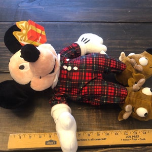 May include: A plush Mickey Mouse wearing red and green plaid pajamas with a brown reindeer plush toy. The Mickey Mouse plush has a red gift box tag that says "Happy Holidays".