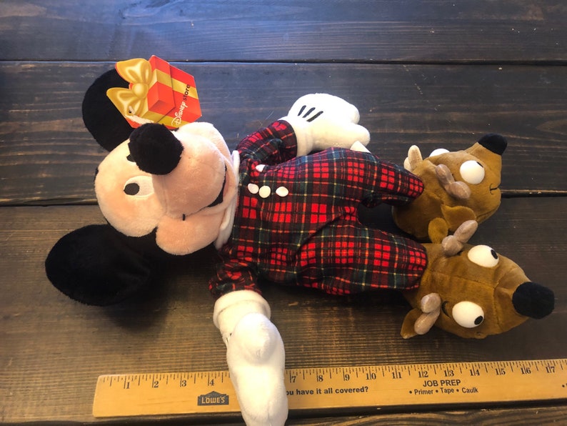 May include: A plush Mickey Mouse wearing red and green plaid pajamas with a brown reindeer plush toy. The Mickey Mouse plush has a red gift box tag that says "Happy Holidays".